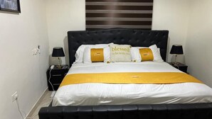 A neatly made bed with a black headboard, white bedding, and a yellow accent. There are two bedside tables with lamps, and a wall-mounted light fixture above the bed. - GHL. G Floor En-suites 2BR Sleep 4. WiFi, DSTV, SOLAR ,AC And Free Breakfast (Kasoa Ghana)