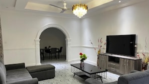 A modern living room with a flat-screen TV, grey sofas, a coffee table with a vase, and a chandelier.