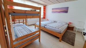 A room with two wooden bunk beds, a single bed, a radiator, a painting on the wall, a chair, and a small table with a lamp.