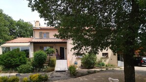 Une maison à deux étages, avec un toit recouvert de tuiles, un porche à l’avant et un petit jardin avec un chemin de gravier menant à l’entrée.