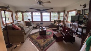 A cozy living room with a sofa, armchairs, a coffee table, and a rug. There are windows with a view of a lake, a ceiling fan, and a television. - Idyllic 3-Bedroom Lake Front Cottage on Ford Lake (Fountain)