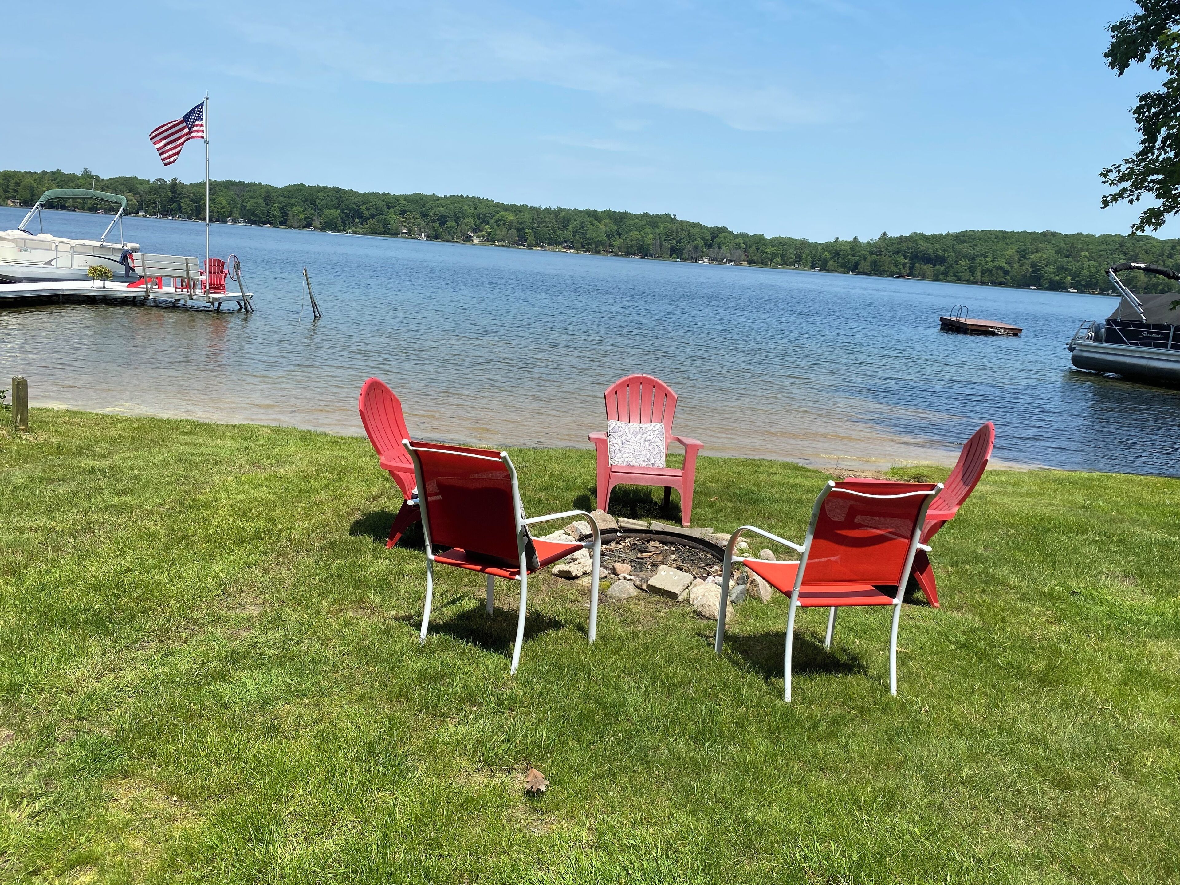 Un paisaje junto al lago con sillones Adirondack rojos alrededor de un brasero, un muelle con un bote y una orilla boscosa.
