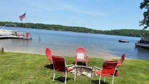 A lakeside setting with red Adirondack chairs around a fire pit, a dock with a boat, and a forested shoreline. - Idyllic 3-Bedroom Lake Front Cottage on Ford Lake (Fountain)