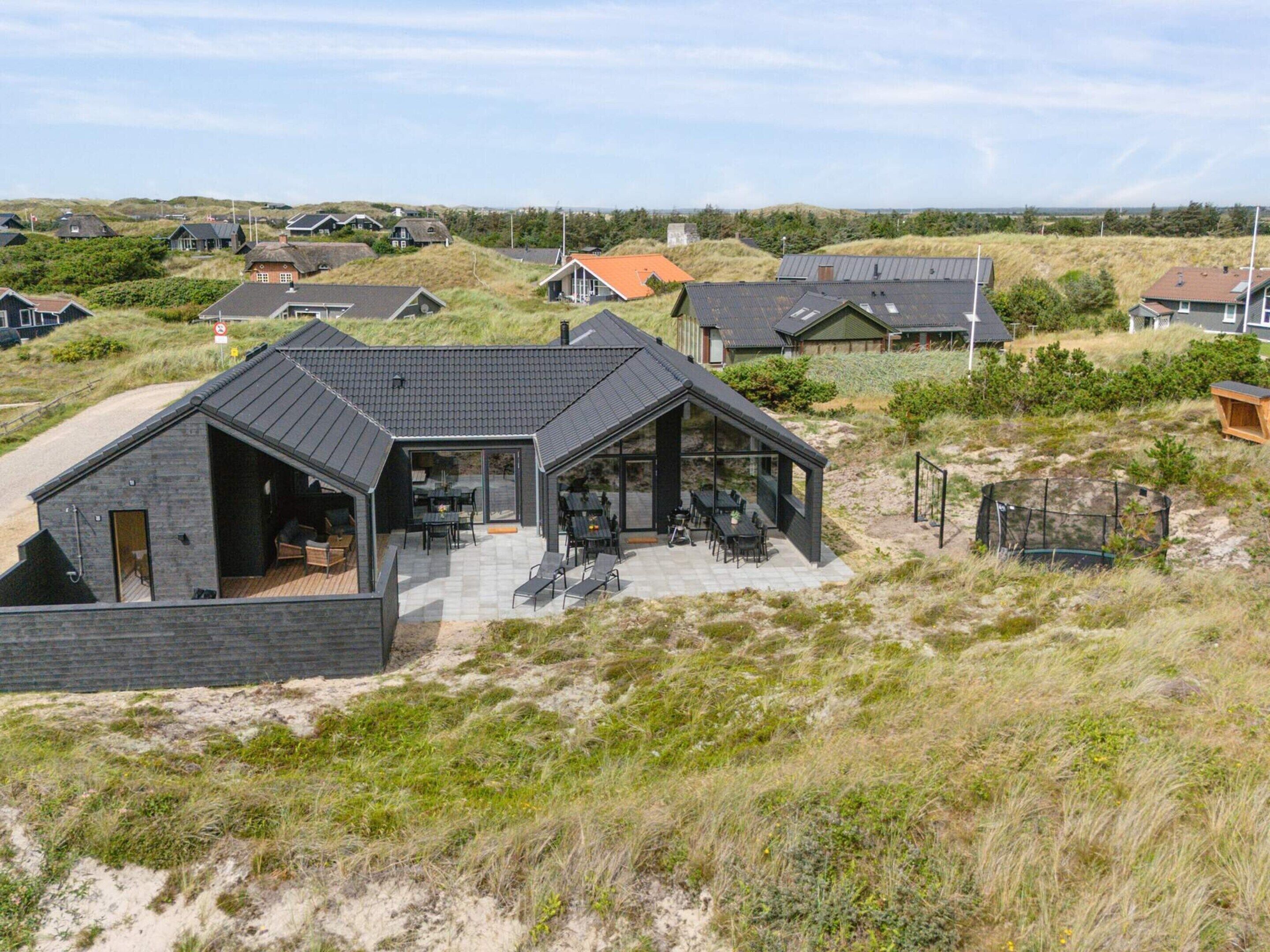 A modern house with a large patio area, surrounded by dunes and other houses.