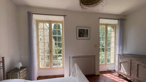 Ein Schlafzimmer mit Bett, Kommode, Heizkörper und zwei mit Vorhängen versehenen Fenstern.