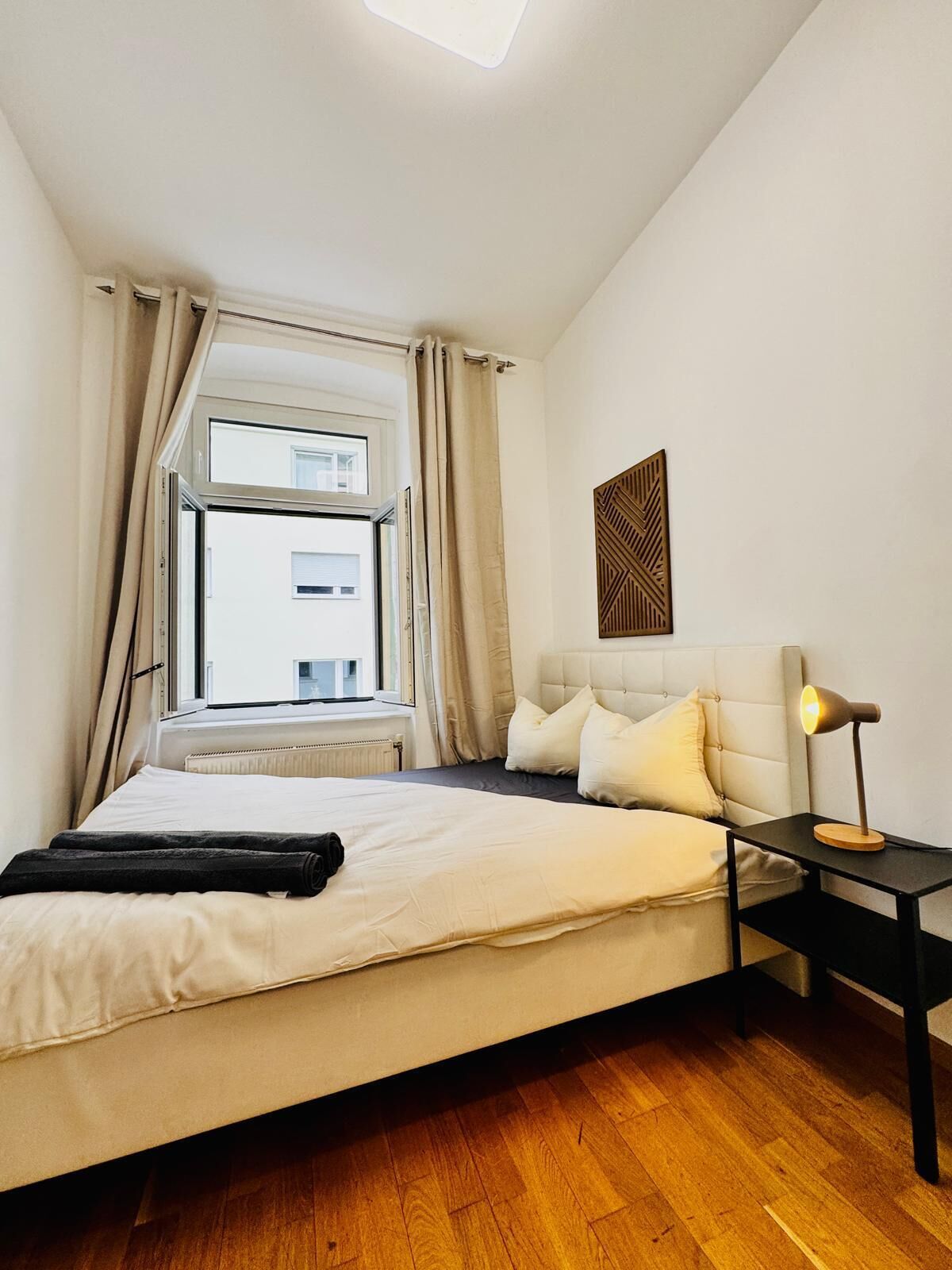A bedroom with a bed, a nightstand with a lamp, a window with curtains, and a wall-mounted artwork.