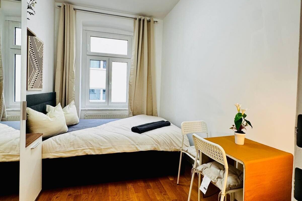 Una habitación pequeña con cama, una mesita con silla, una planta en maceta y un ventanal con cortinas.