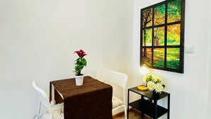 Een kleine kamer met een donkerbruine tafel, een witte stoel, een klein wit tafeltje en een schilderij aan de muur.