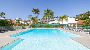 A rectangular swimming pool with clear blue water, surrounded by a paved deck. There are lounge chairs and umbrellas along the poolside.