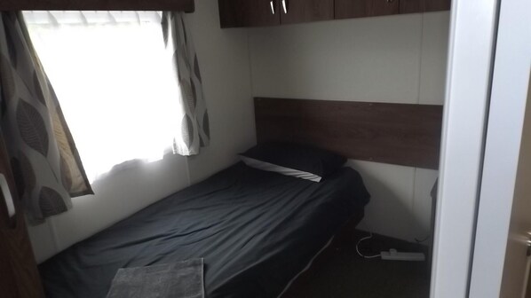 A small, single-bed room with a wooden headboard, a window with sheer curtains, and a power outlet on the floor. - Modern 3 Bedroom Lodge at Avonvale Holiday Lodges (Evesham)