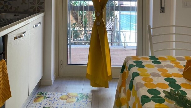 Una habitación pequeña y luminosa con una cocina, un balcón con cortina amarilla y una cama con edredón de lunares.