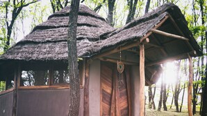 A rustic cabin with a thatched roof, wooden walls, and a stone foundation. It features a wooden door, a small porch, and a hanging dreamcatcher.