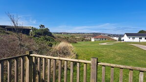 A grassy area with a wooden fence, a white building with a red roof, and a hill in the background. - D48 Sunvale, Riviere Towans (Hayle)