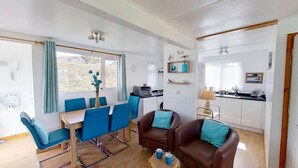 A compact living space with a dining area, a kitchenette, and a seating area with a view of greenery outside. - D48 Sunvale, Riviere Towans (Hayle)