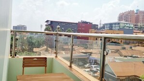 A balcony with a wooden bench, glass railing, and a view of surrounding buildings. - BLT Suites - Sunset view (Kiira)