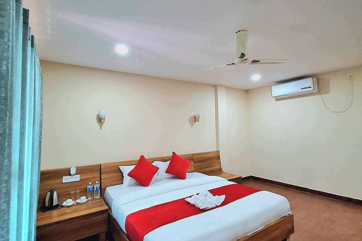 A hotel room with a wooden bed, red and white bedding, a nightstand with a kettle and glasses, and an air conditioning unit on the wall.