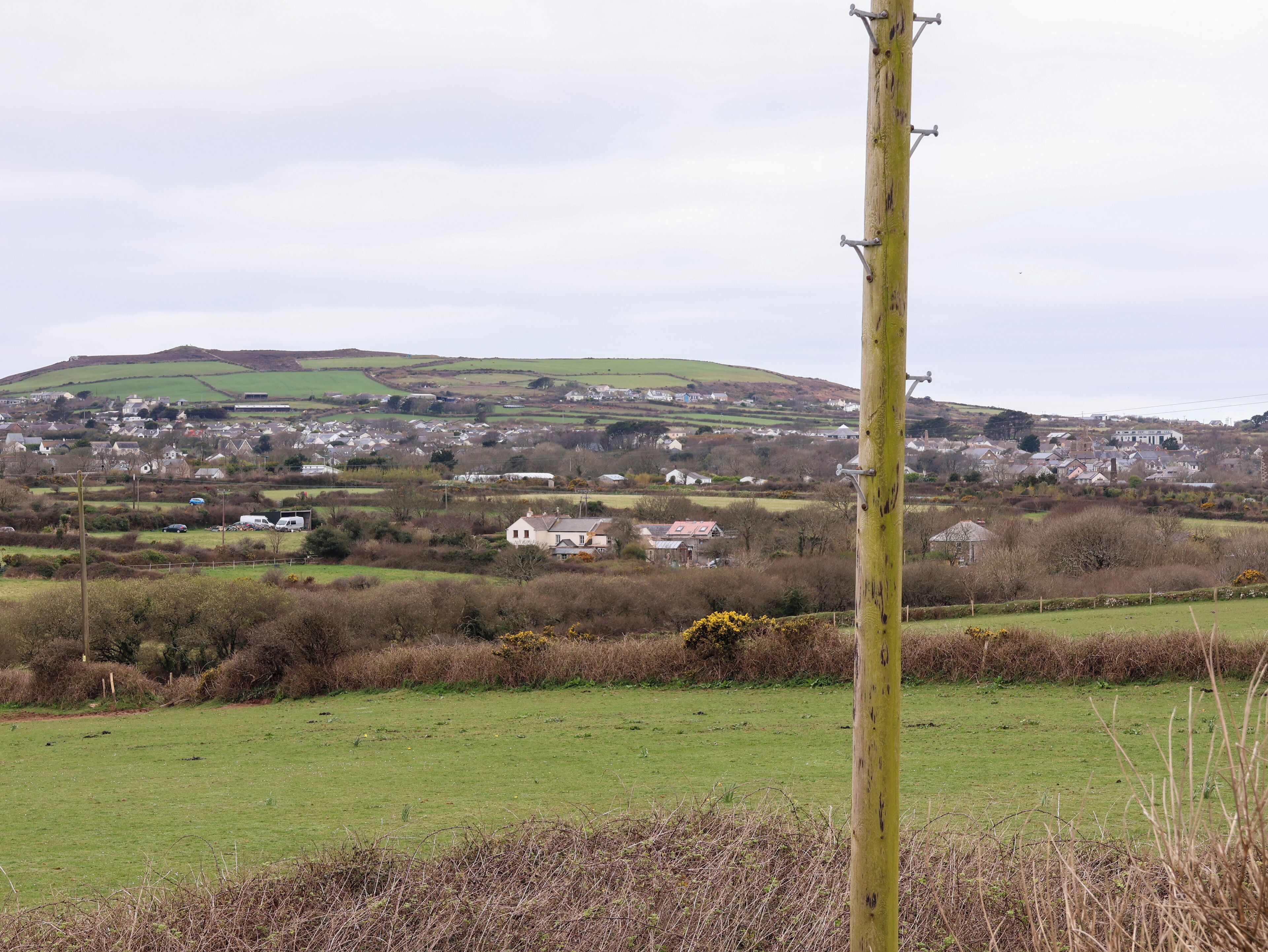 A rural landscape with a village in the background, a prominent utility pole in the foreground, and a green field in the middle ground.