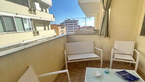 Un balcon avec des chaises blanches et une table, un livre, et une vue sur des bâtiments en arrière-plan.