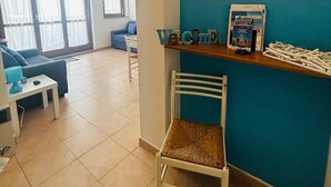 Un salon avec un canapé bleu, un fauteuil blanc et une petite table sur laquelle se trouve une lampe. Un mur bleu est orné d’une étagère où sont posés des livres et d’un tableau encadré.