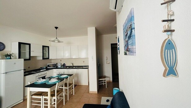 Une cuisine de taille réduite avec des meubles blancs, un coin repas doté d’une table noire et de chaises blanches, et une décoration représentant un poisson bleu sur le mur.