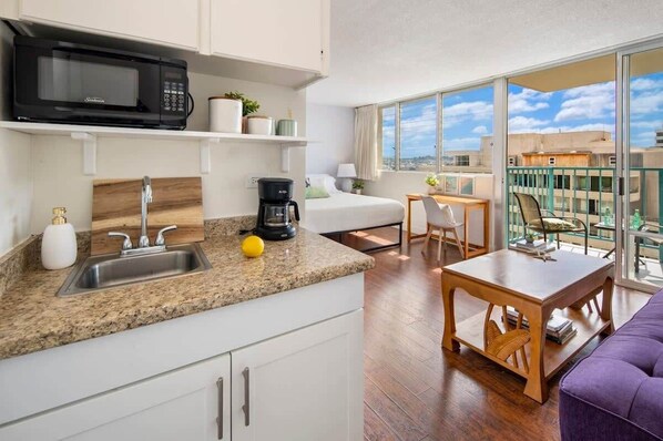 A compact kitchen with a microwave, sink, and coffee maker, a balcony with a table and chairs, and a view of the cityscape. - @ Marbella Lane - Marina Studio Near the Beach (Honolulu)