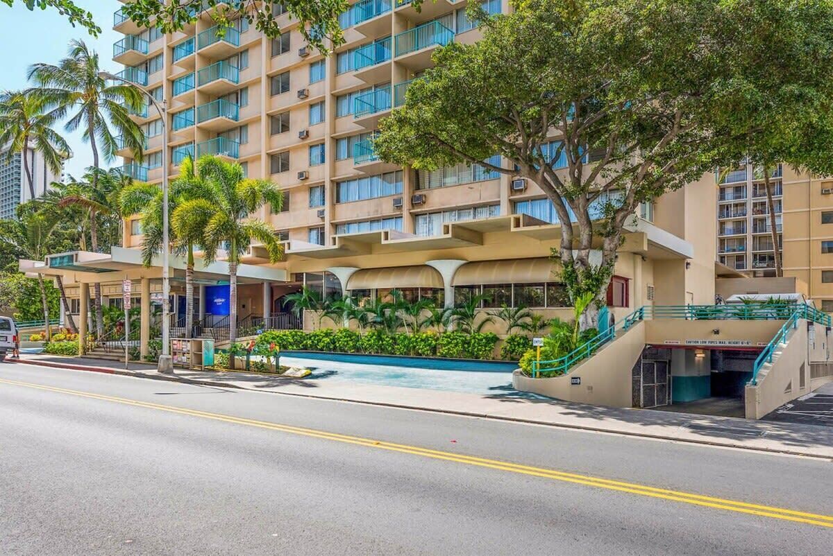 A multi-story hotel with a pool area, palm trees, and a staircase leading to the entrance.