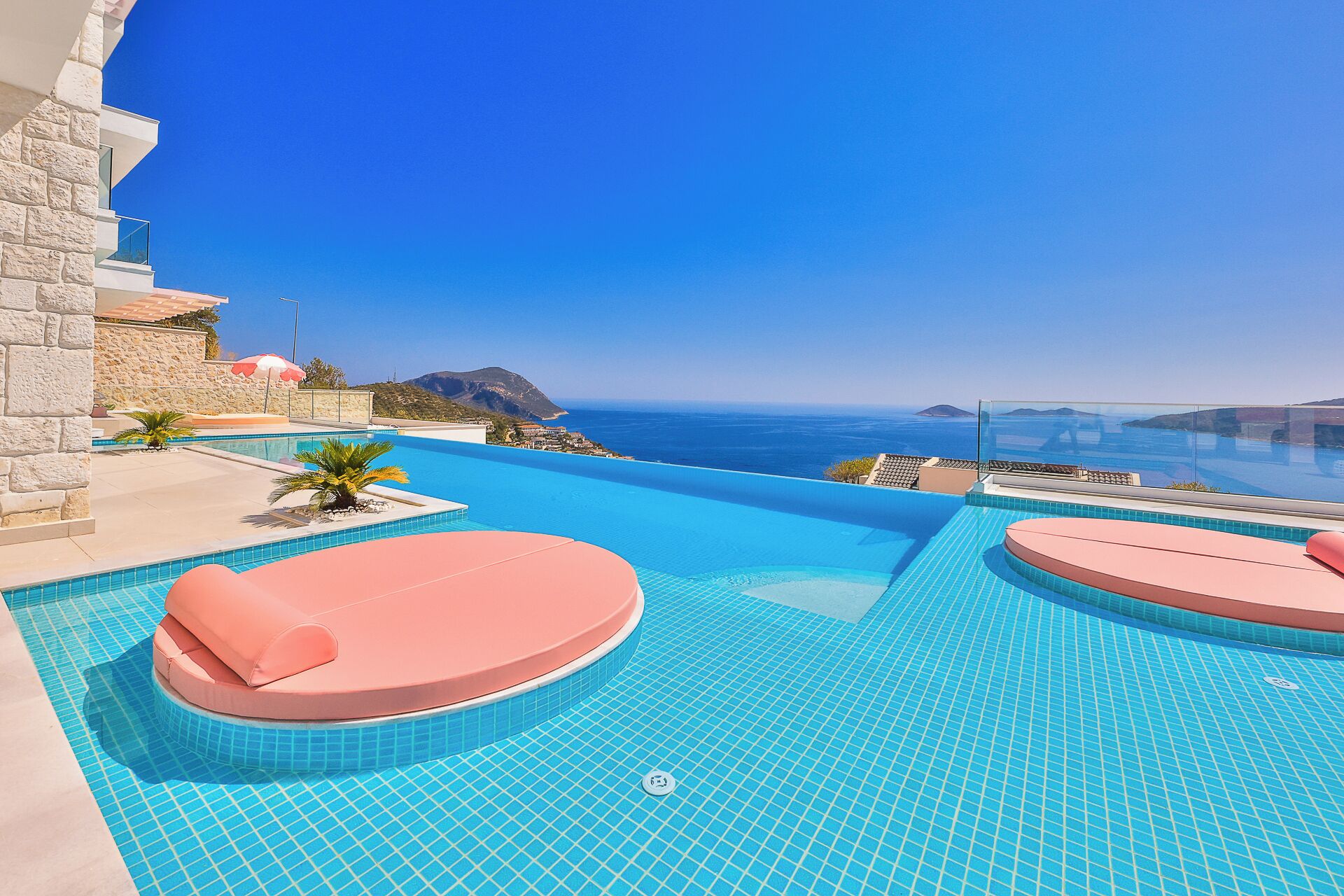 Una piscina infinita con tumbonas rosadas, una pared de piedra y vistas al mar.