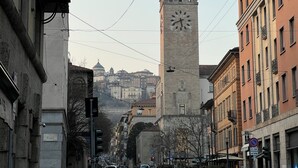 Una calle urbana con una imponente torre de reloj, edificios con balcones y un cielo despejado.