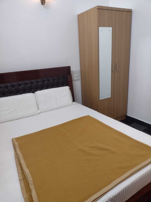 Un lit bien fait, recouvert d’une couverture marron, une armoire en bois avec un miroir, et une prise électrique au mur.