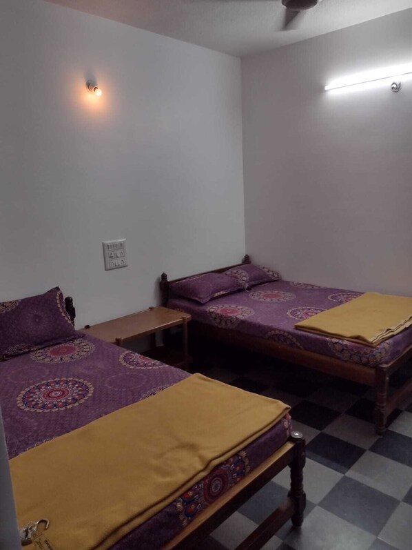A room with two beds, each with a patterned bedspread and a yellow blanket. There is a small wooden nightstand between the beds, and a wall-mounted light fixture above the left bed. - Padmashree lodge (Mudigere)