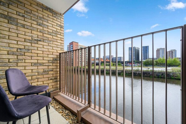 A balcony with a brick wall, two dark chairs, and a metal railing overlooking a river with buildings in the background. - Manchester - Apartment - Sleeps 5 - Parking (Salford)