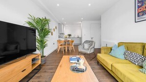 A modern living room with a flat-screen TV, a yellow sofa, a wooden coffee table, and a dining area in the background. - Manchester - Apartment - Sleeps 5 - Parking (Salford)