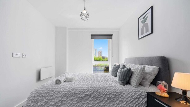 A modern bedroom with a large bed, a nightstand, a lamp, a geometric light fixture, a framed picture, and a window with a city view. - Manchester - Apartment - Sleeps 5 - Parking (Salford)