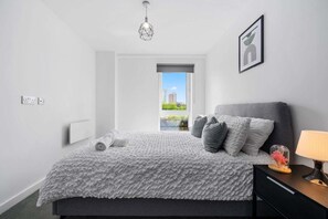 A modern bedroom with a large bed, a nightstand, a lamp, a geometric light fixture, a framed picture, and a window with a city view. - Manchester - Apartment - Sleeps 5 - Parking (Salford)