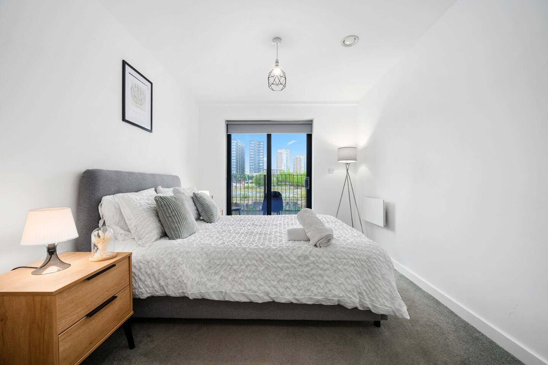 Quarto moderno com cama, mesa de cabeceira, abajur e vista para prédios pela janela.