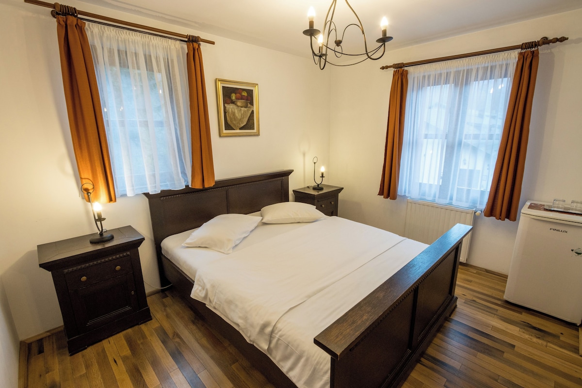 A bedroom with a wooden bed, a nightstand, a bedside table with a lamp, a refrigerator, and a window with curtains.