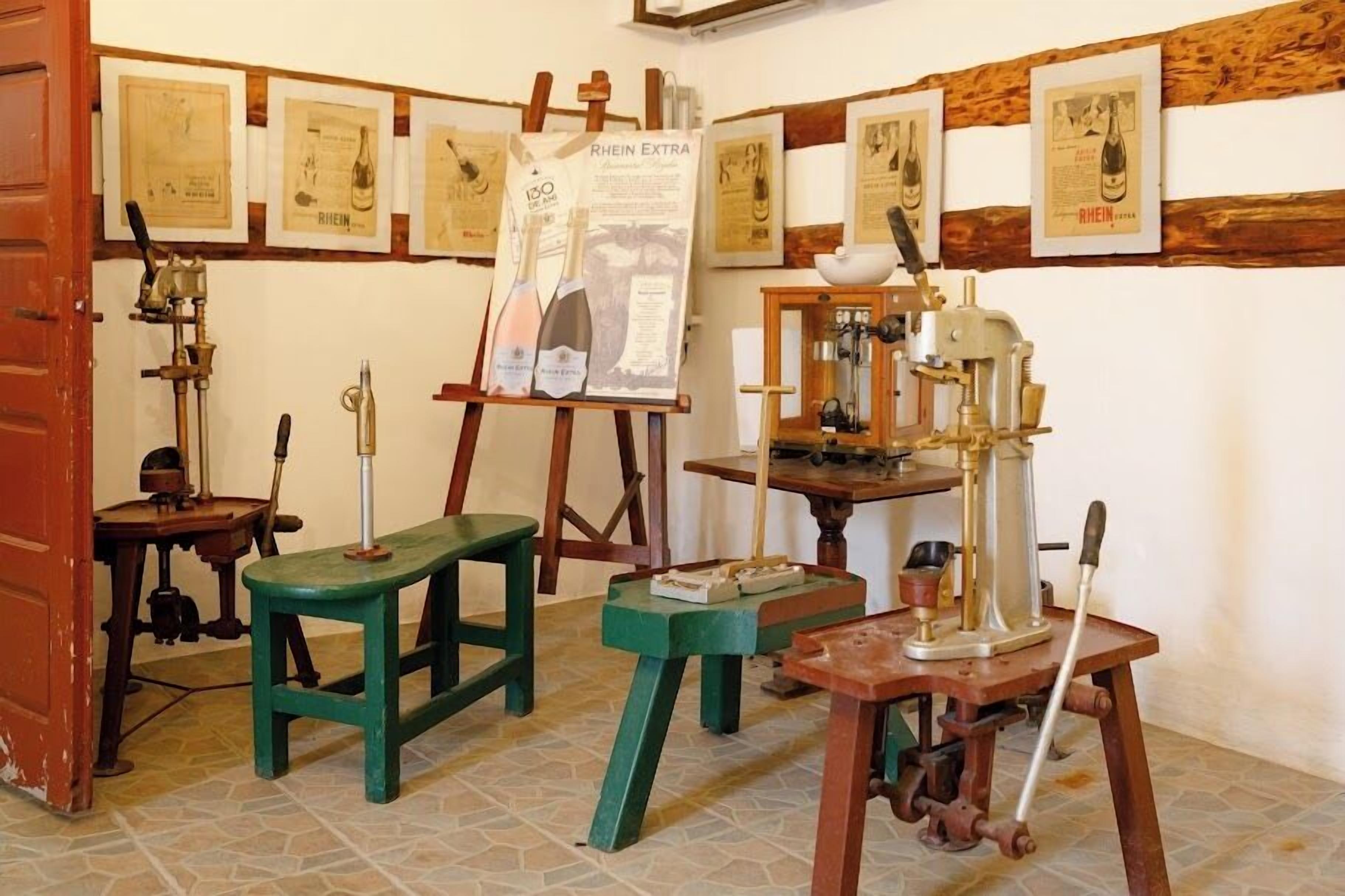 A room with vintage wine-making equipment, green stools, and framed posters on the wall.