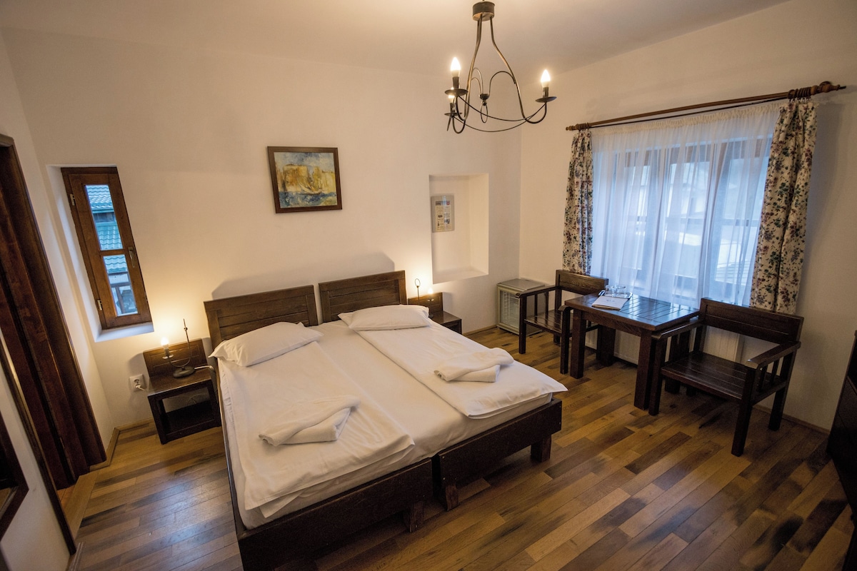 A bedroom with a double bed, bedside tables, a desk with a chair, a chandelier, and a window with curtains.