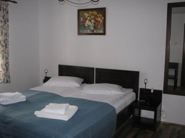 A neatly made bed with white linens and a blue blanket, a bedside table with a lamp, and a painting of flowers on the wall. - Pensiunea Rhein (Azuga)