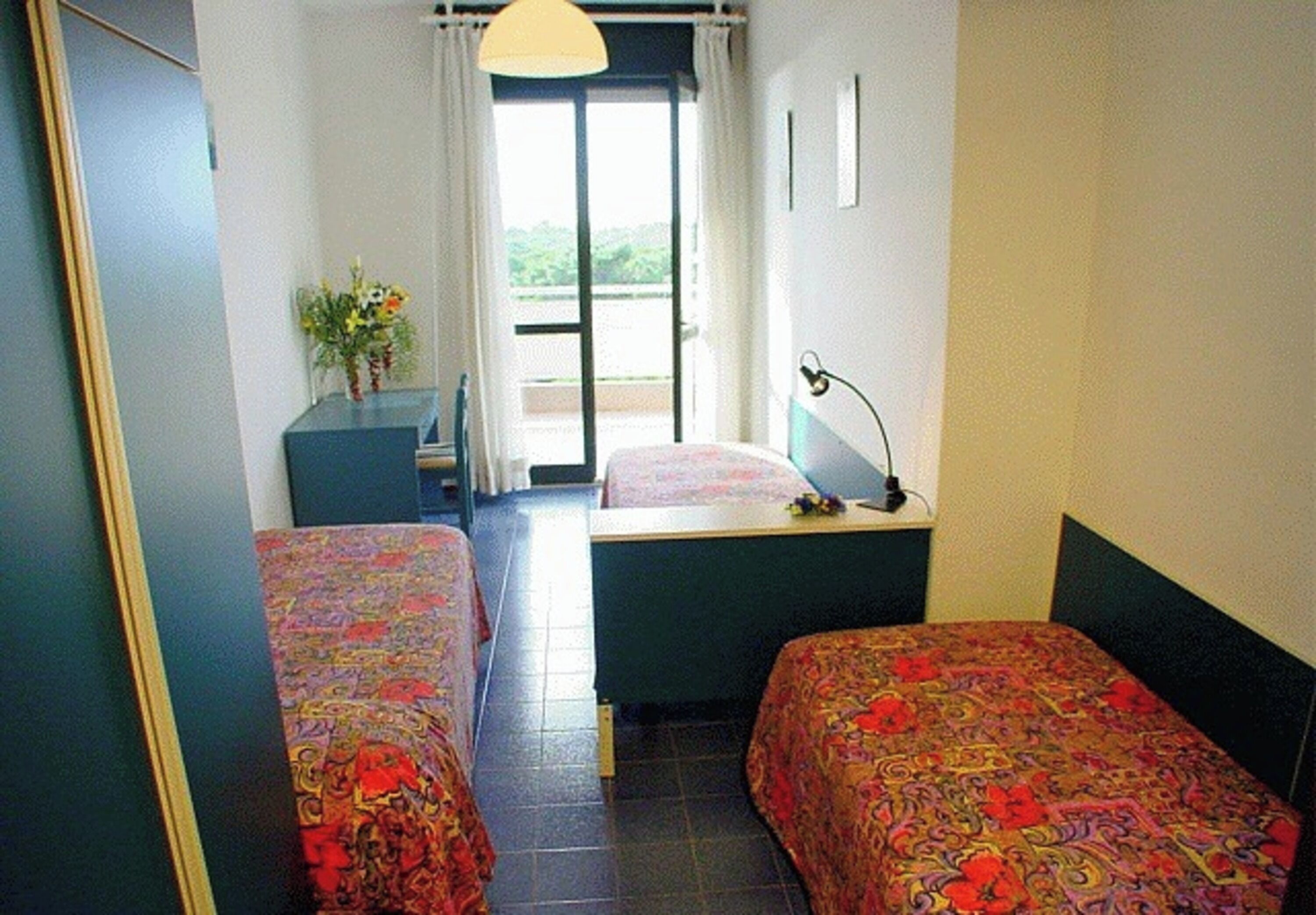 A room with two beds, a desk, a chair, a lamp, and a window with curtains.