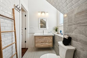 A bathroom with a white tiled wall, a wooden vanity, a white sink, a mirror, and a toilet.