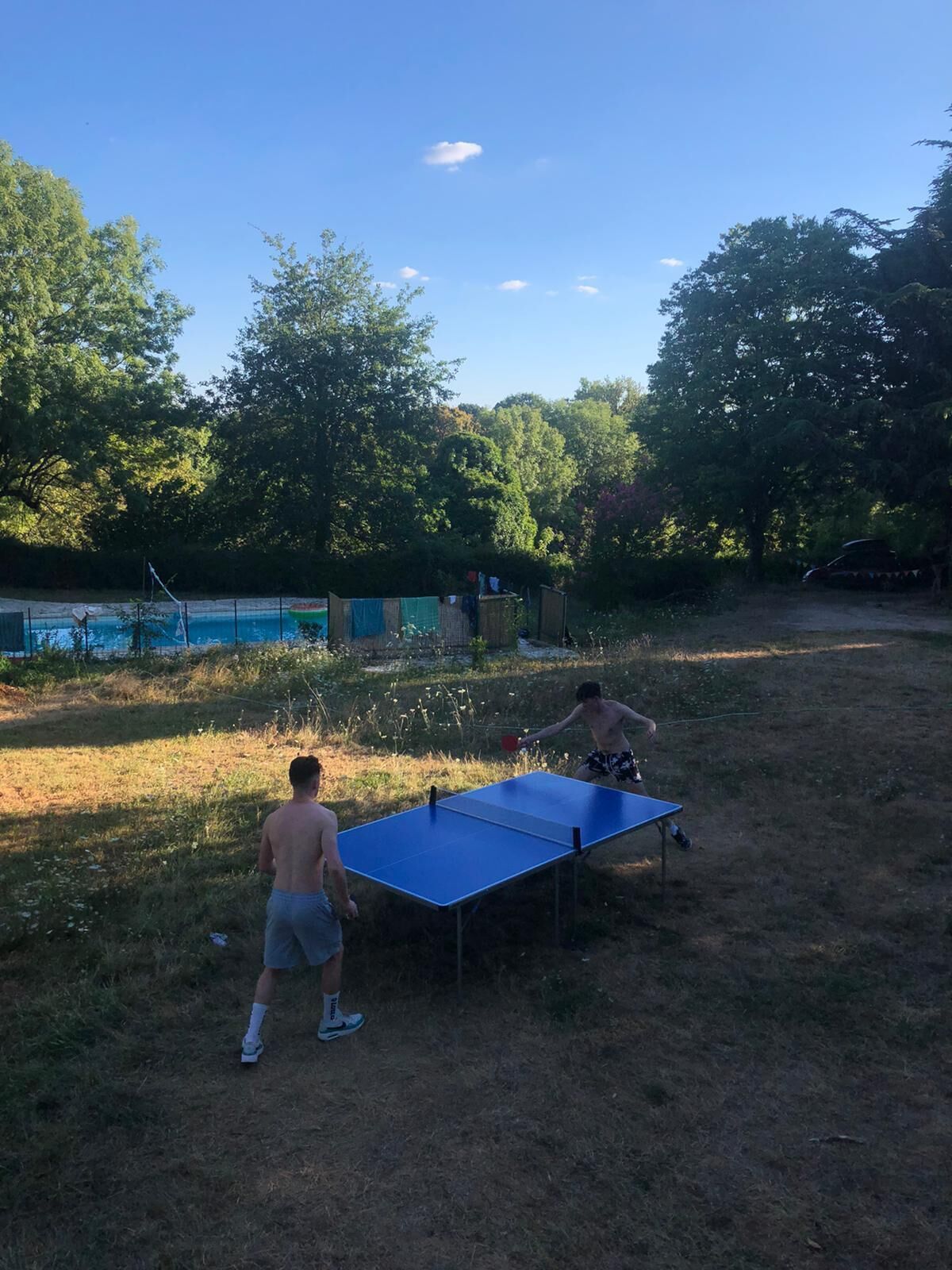 Zwei Personen spielen Tischtennis im Freien, im Hintergrund ist ein Schwimmbecken zu sehen.