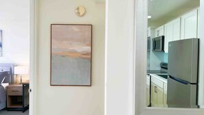 Una habitación con un cuadro en la pared, una cama, una mesita de noche y vistas a una cocina con refrigerador y microondas.