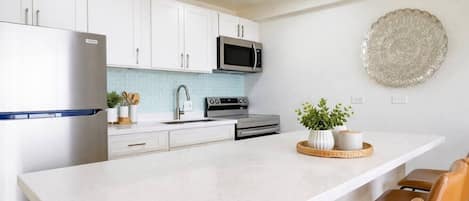 A modern kitchen with white cabinetry, a stainless steel refrigerator, a microwave, and a stove. There is a large island with a sink and a potted plant.