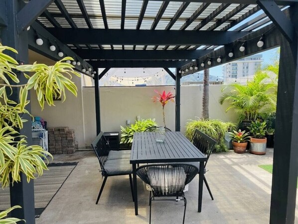 A covered patio area with a table and chairs, surrounded by potted plants and a view of buildings.