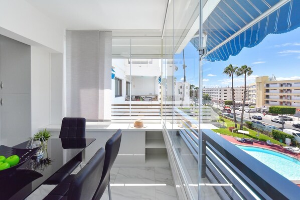 A balcony with a glass railing, a dining area with a table and chairs, and a view of a swimming pool and buildings in the background. - Iguazu 304 by VillaGranCanaria (Playa del Ingles)