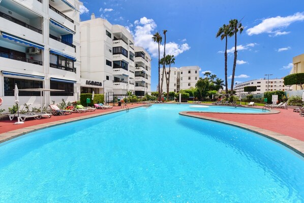 Una piscina con sillones de descanso y palmeras, rodeada de edificios de varios pisos con balcones.