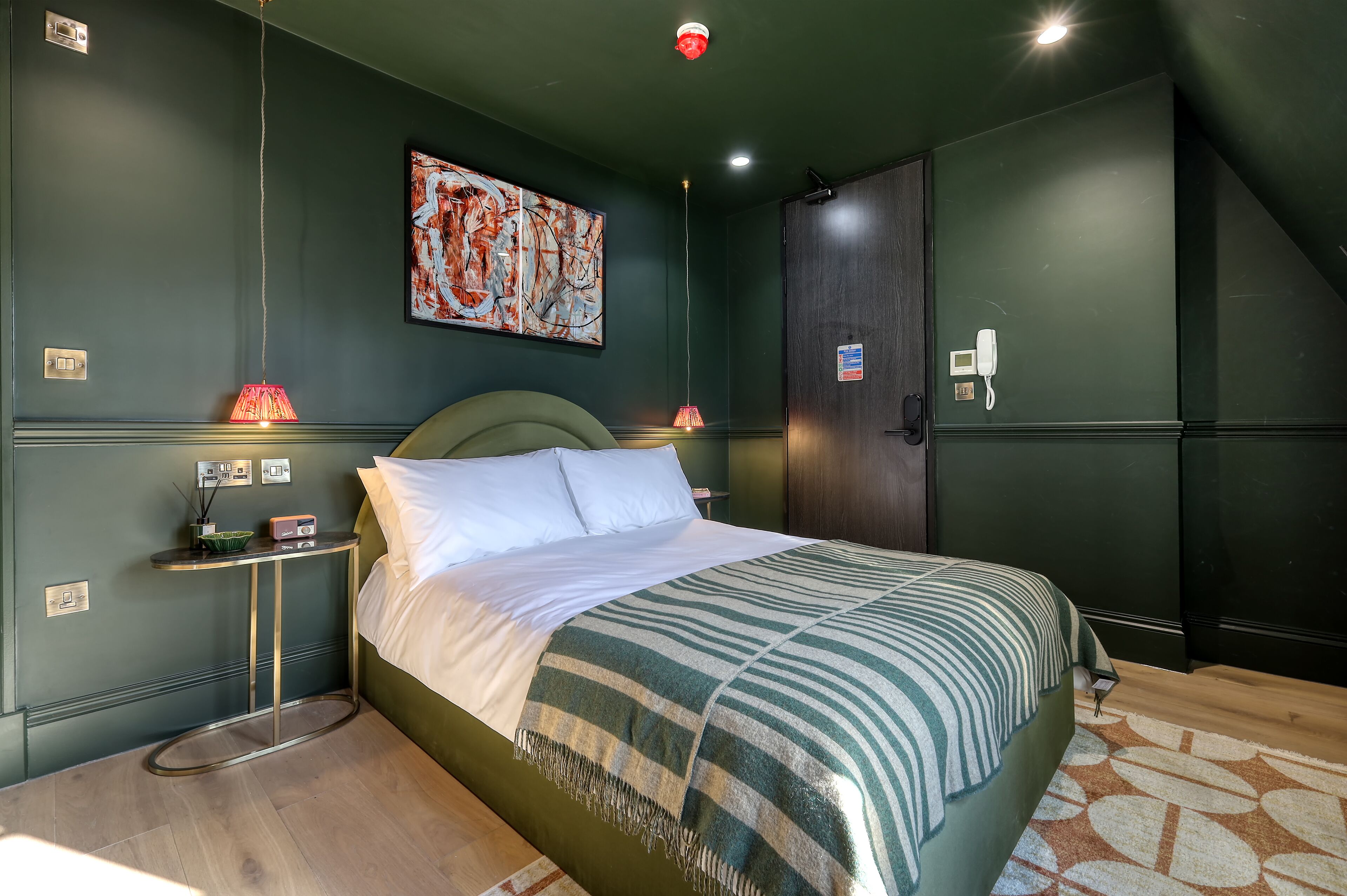 A neatly made bed with a striped blanket, a bedside table with a lamp, a wall-mounted painting, and a phone on the wall.