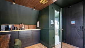 A modern kitchen with dark green walls, wooden ceiling, and a wooden floor. There is a bar area with a green chair and a coffee machine. - The Carlton Tavern Hotel (London)