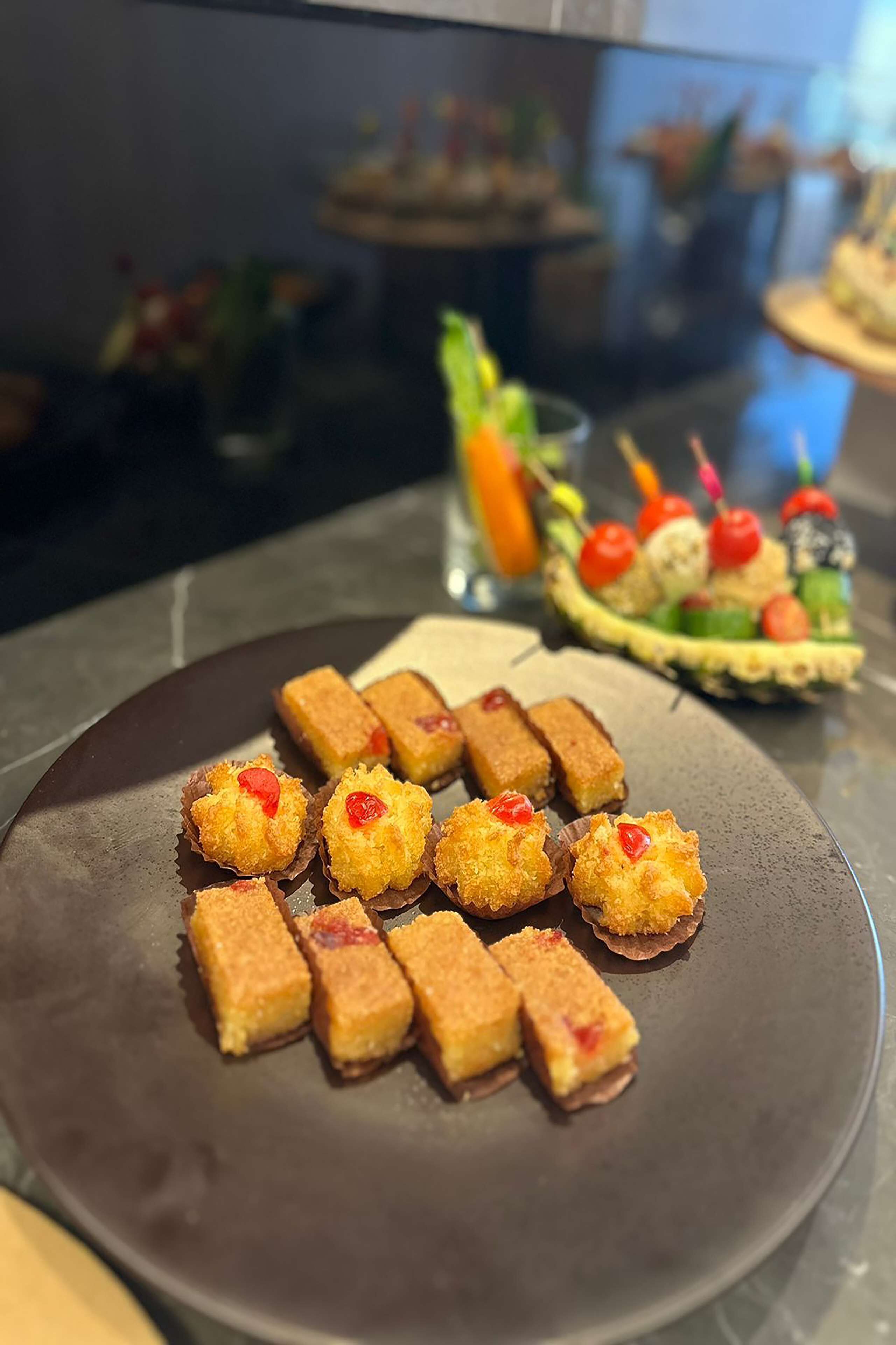 Ein Teller mit Speisen, darunter kleine, rechteckige Gebäcke mit roter Frucht, sowie ein Dessert mit verschiedenen Früchten und einer Kirsche.
